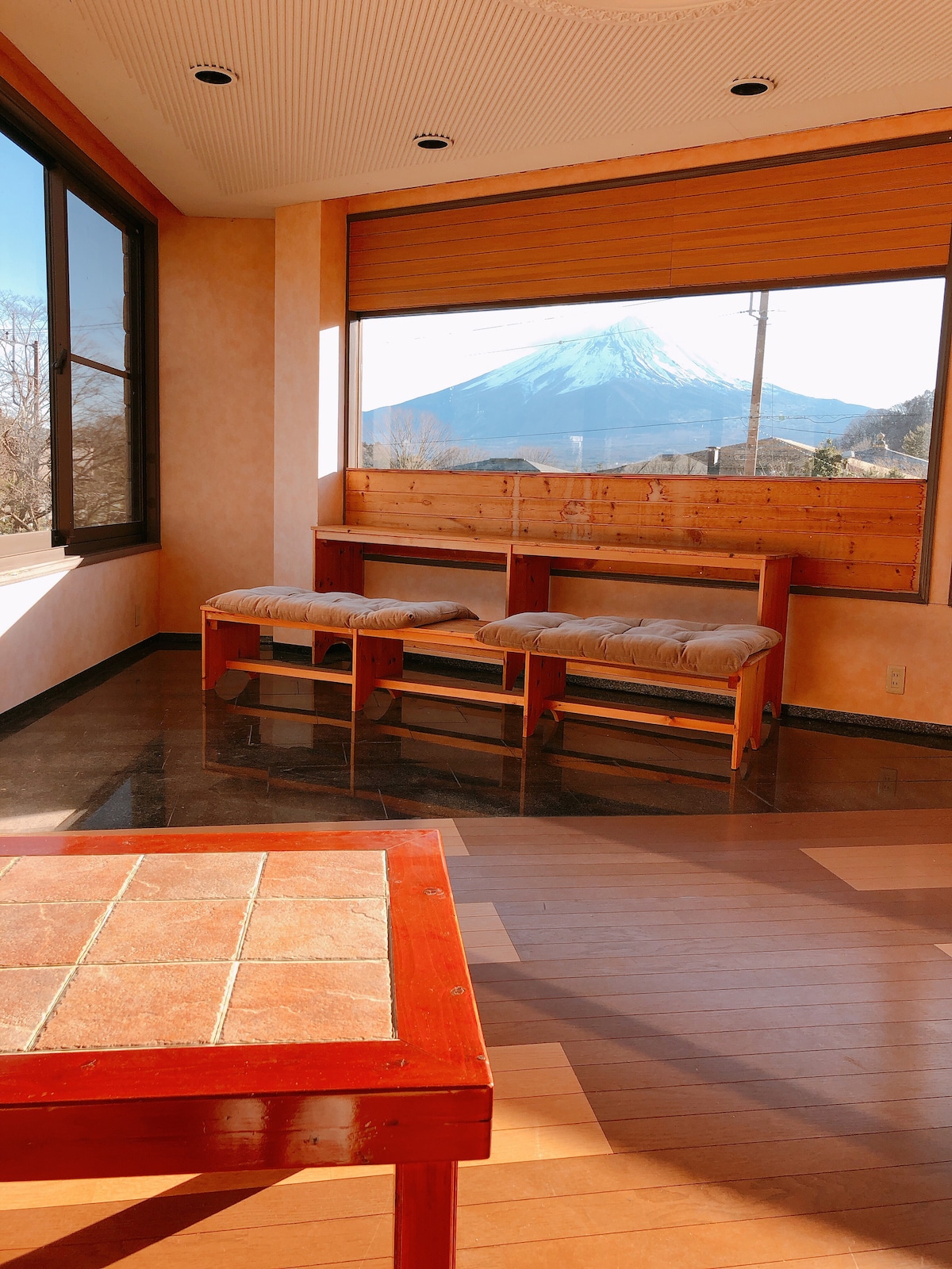 A spacious lounge area features large windows revealing a view of Mount Fuji. Two cushioned benches are positioned against the window, and a wooden table is seen in the foreground, enhancing the serene atmosphere of the space.