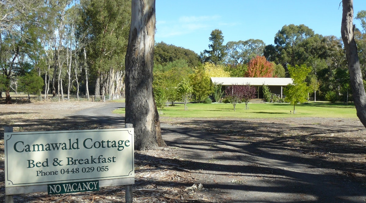 A sign marking Camawald Cottage is positioned at the entrance, with the cottage set back among a variety of trees and landscaped areas. A gravel path leads to the cottage, which features a covered porch surrounded by lush greenery and colorful seasonal foliage.