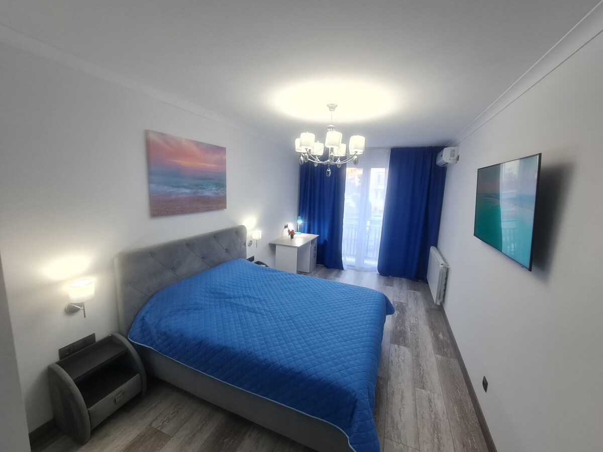 A spacious bedroom is fitted with a large, soft bed covered in a blue quilt. Two bedside lamps provide gentle lighting, while blue curtains frame a balcony door. A modern chandelier hangs from the ceiling, and a wall-mounted television adds a contemporary touch.