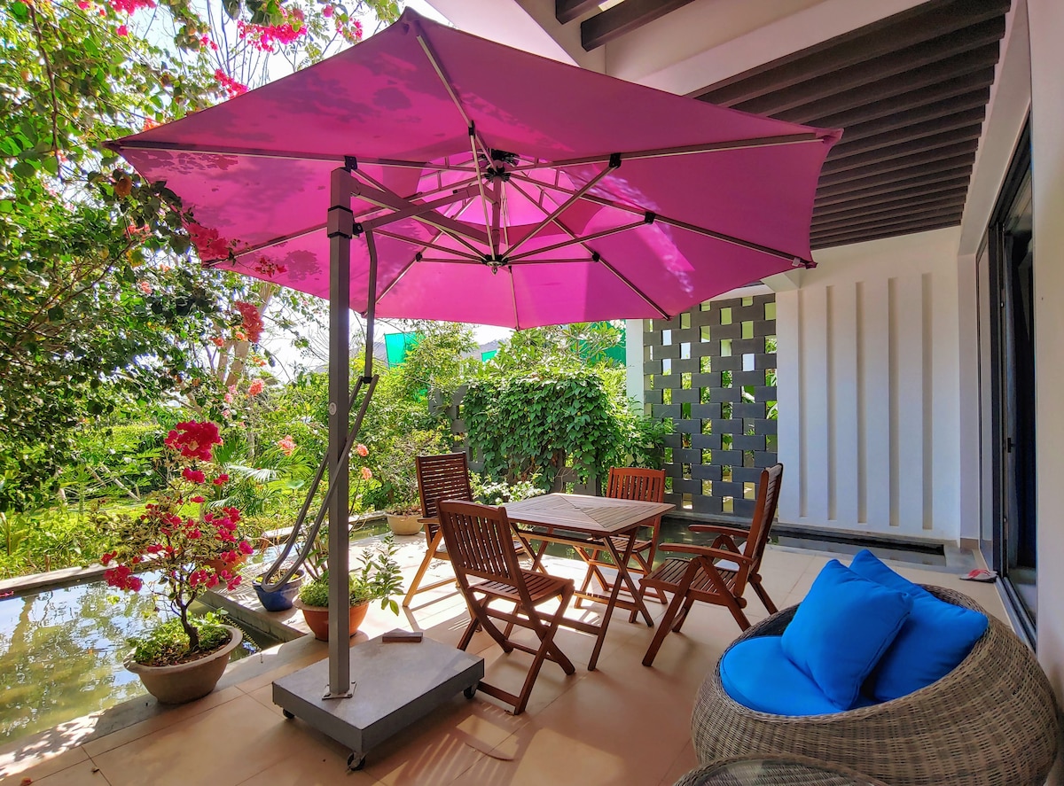 An outdoor garden patio features a light purple umbrella shading a wooden table surrounded by four folding chairs. Potted plants and vibrant flowers add natural beauty, while a comfortable blue cushion is placed on a nearby lounge chair. Lush greenery and a decorative wall provide a serene backdrop.