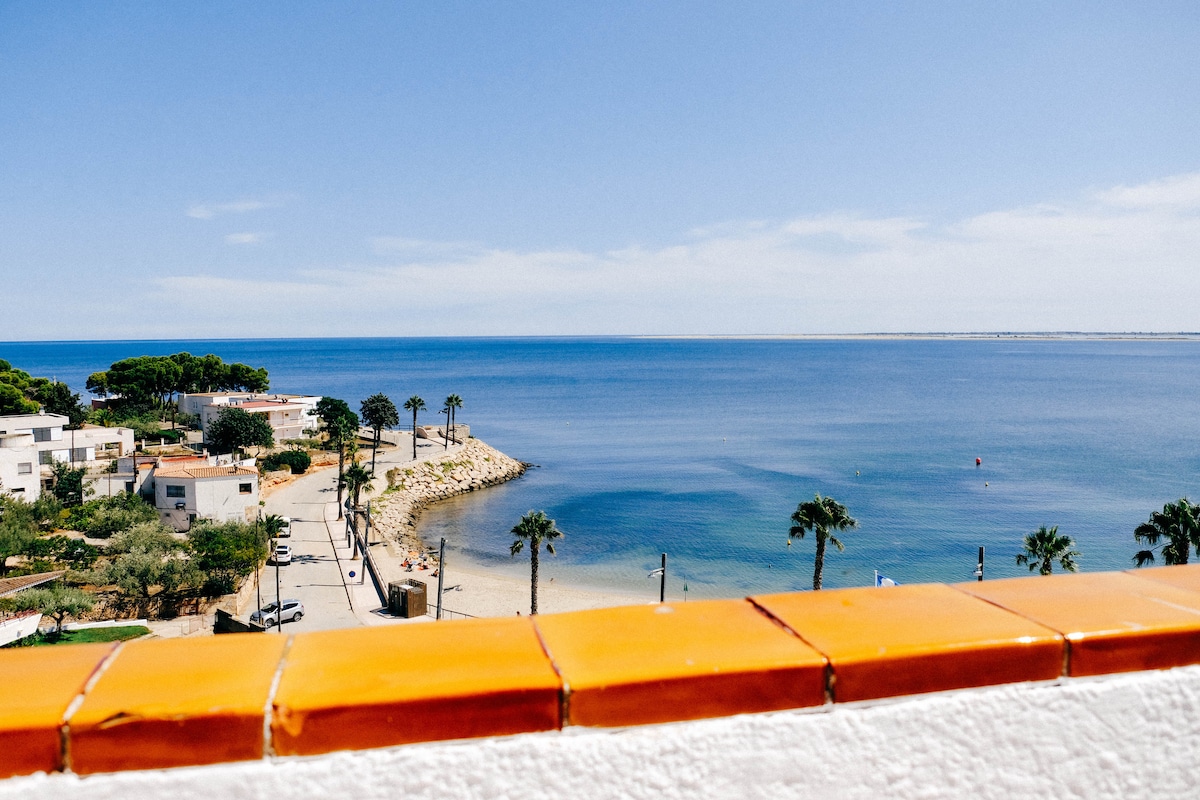 A panoramic view showcases the serene coastline along the blue waters, framed by green palm trees and sandy beaches. The image captures the peaceful atmosphere of the area, highlighting nearby residential buildings and walking paths under a clear sky.