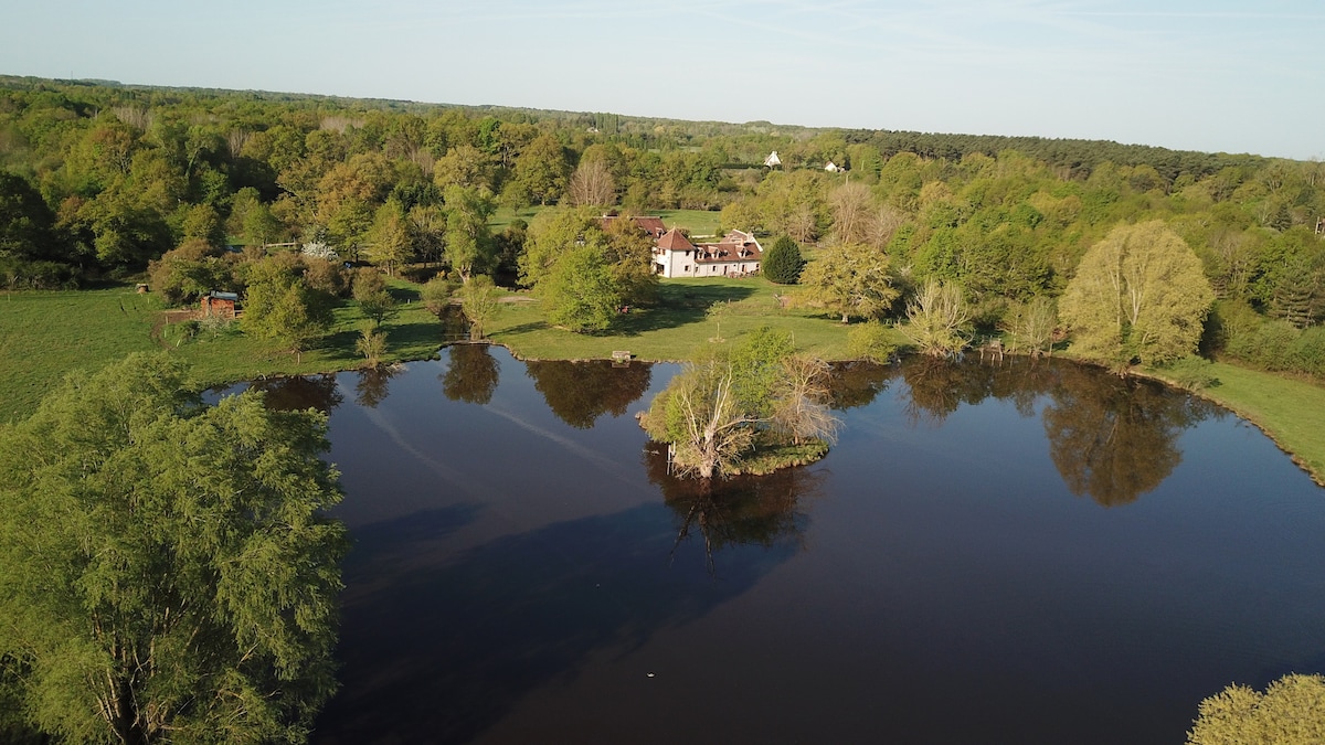 An aerial view captures a serene landscape, featuring a tranquil pond surrounded by lush greenery. A charming building is positioned in the background, framed by trees and open fields, reflecting the natural beauty of the area.