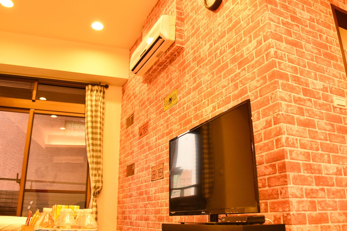 A cozy living area is presented with an exposed brick wall, creating a warm and inviting ambiance. A sleek television is positioned in front, while natural light filters through large windows dressed in checkered curtains, offering a view of the bustling surroundings.