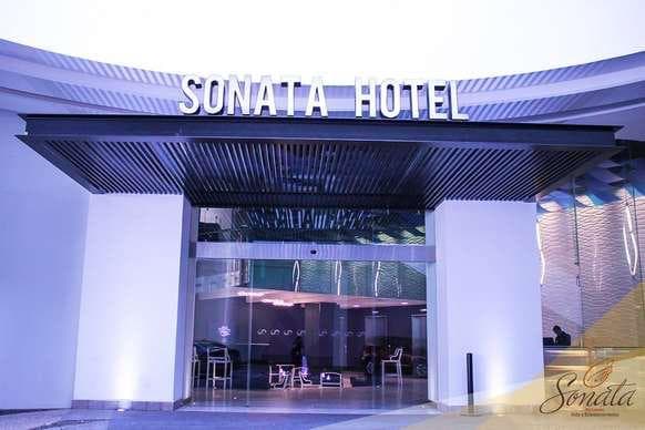 The hotel exterior is characterized by a modern design, featuring a sleek façade and large glass doors that provide an inviting entryway. The name 'Sonata Hotel' is prominently displayed above, framed by a stylish overhang.