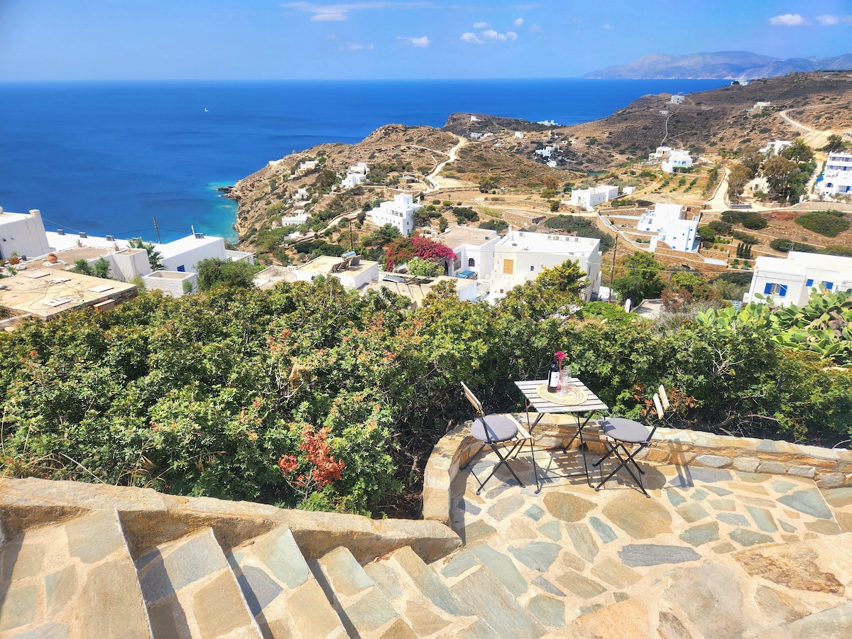 A private terrace offers a scenic view of the Aegean Sea and the village of Chora, framed by lush greenery. A small table with two chairs is positioned on a stone pathway leading down from the property, enhancing the outdoor experience.