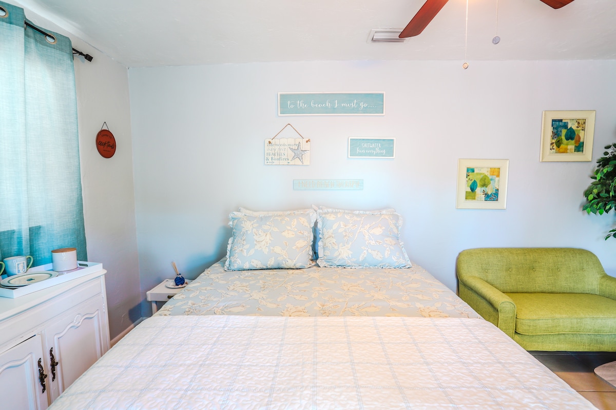 A spacious sleeping area features a queen-size bed with a light, floral-patterned comforter. A soft green chair adds a pop of color next to the bed. Wall decorations include framed art and a decorative sign, enhancing a tranquil ambiance.