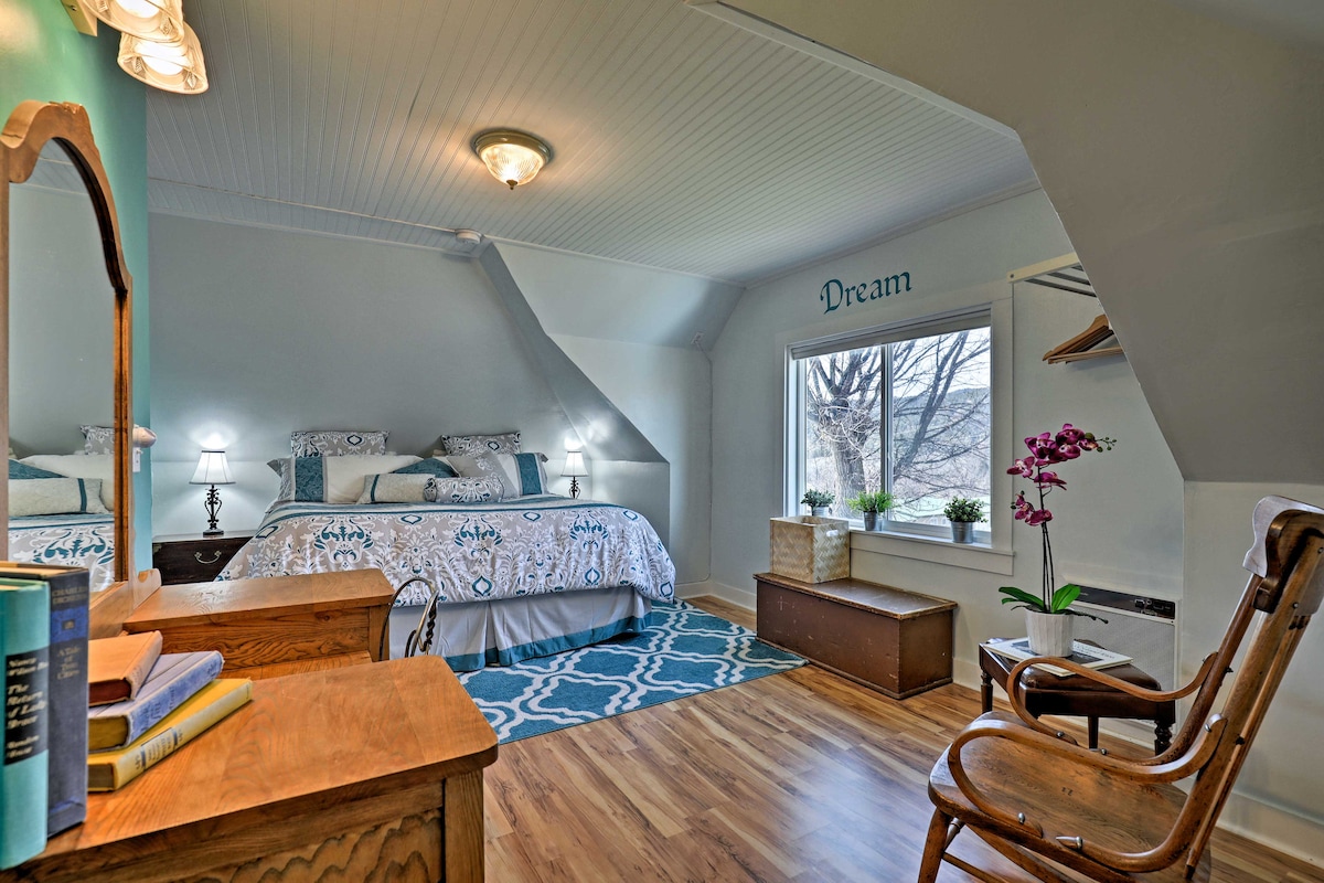 The cozy bedroom features a queen bed adorned with a decorative blanket, flanked by two bedside lamps. A large window allows natural light to illuminate the space, complemented by a small plant on the sill. A wooden dresser and a chair add comfort to the room.