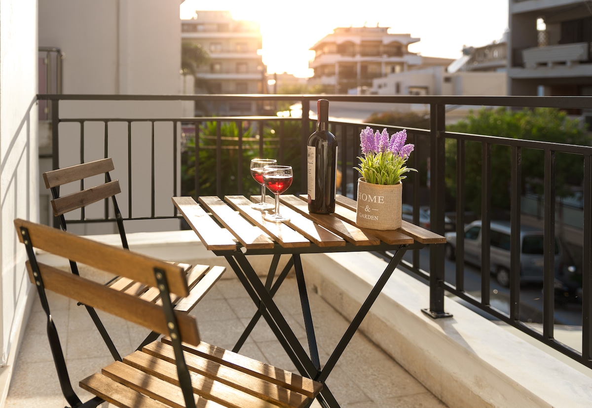 A small balcony is equipped with a wooden table and two folding chairs. A bottle of wine and two glasses are placed on the table, accompanied by a floral arrangement in a pot. Sunlight shines in the background, highlighting the nearby buildings.