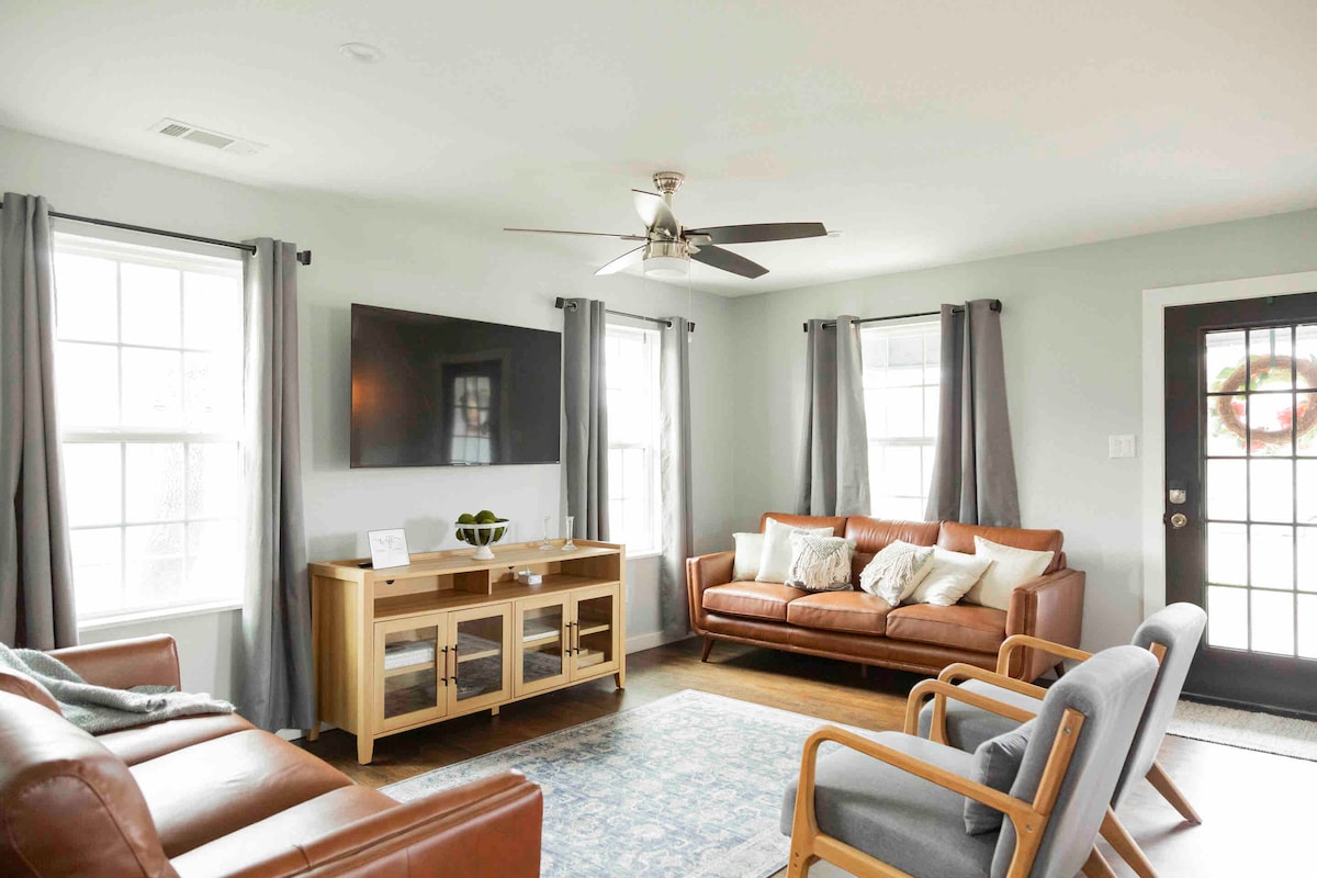 A welcoming living area features a mix of comfortable seating, including a leather sofa and accent chairs. Natural light enters through two large windows, framed with light drapes. A wooden TV stand holds a flat-screen television, adding to the modern design.
