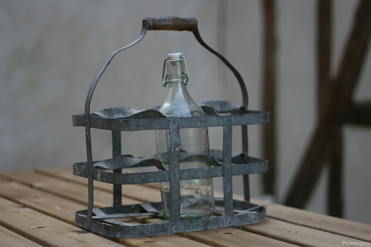 An antique-style metal basket is displayed on a wooden table, designed to hold multiple bottles. A single glass bottle is placed inside, showcasing the craftsmanship of the container. The rustic materials contribute to a classic aesthetic.
