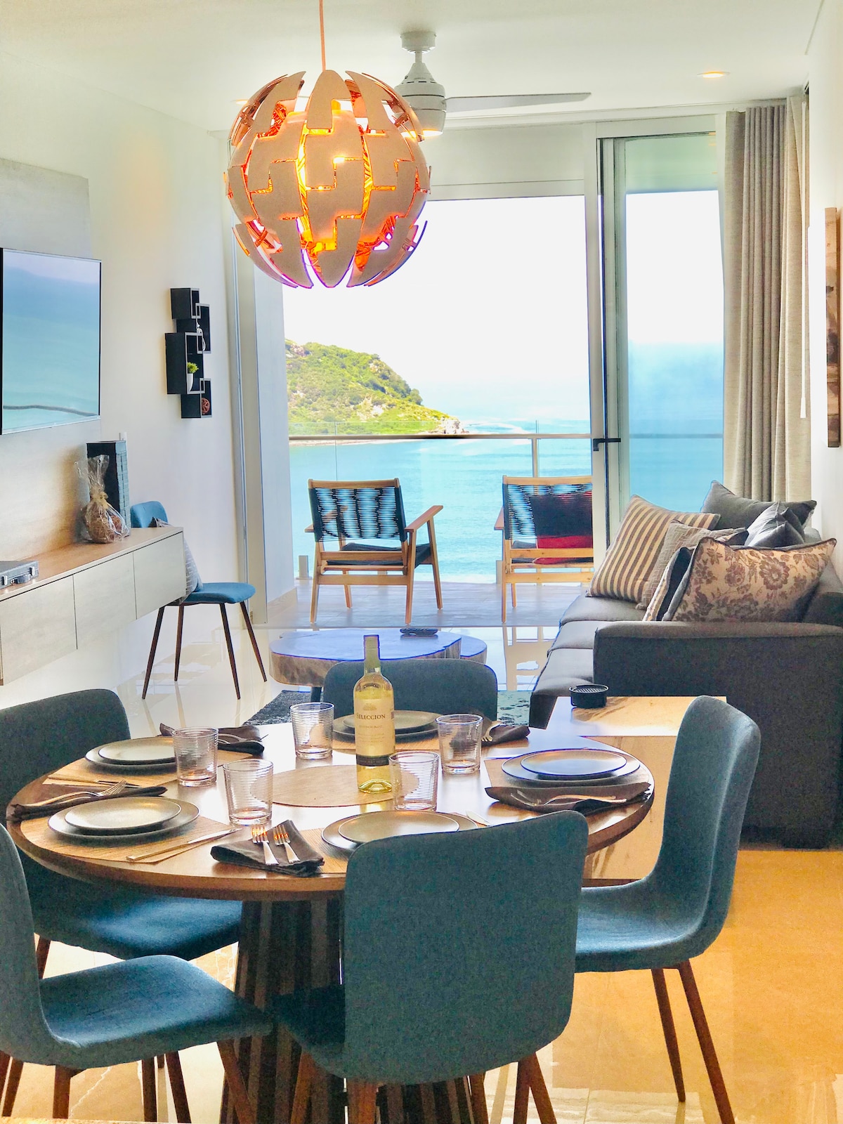 A dining area is featured with a round wooden table set for six, accompanied by blue chairs. A spacious living area with a sofa and armchairs provides a view of the ocean through floor-to-ceiling windows. A unique light fixture hangs above the table.