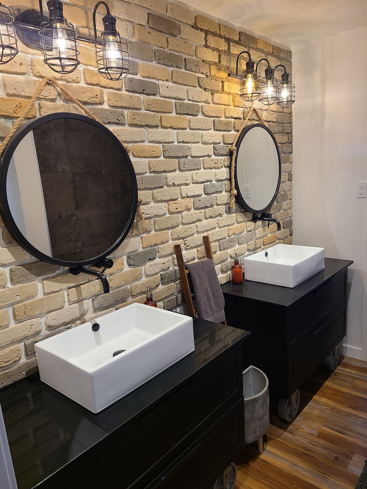 A stylish bathroom space features two circular mirrors above modern rectangular sinks. The backsplash showcases exposed brick, while two industrial-style light fixtures illuminate the area. Black vanities with wheels provide storage beneath the sinks, creating a contemporary, functional environment.