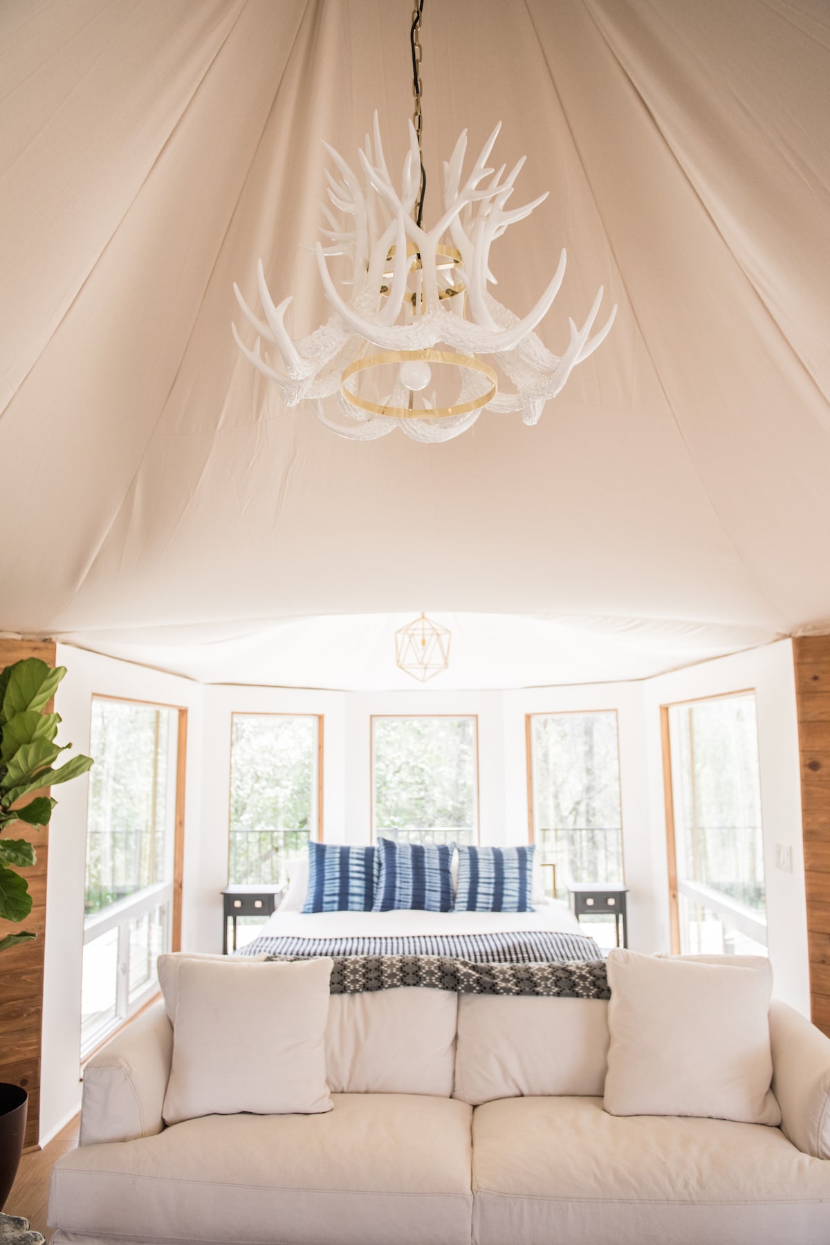 A spacious living area features a cozy sofa adorned with plush white cushions. A unique antler chandelier hangs from the ceiling, offering warm illumination. Large windows allow natural light to fill the space, complemented by a stylish bed with a blue and white throw, positioned in the background.