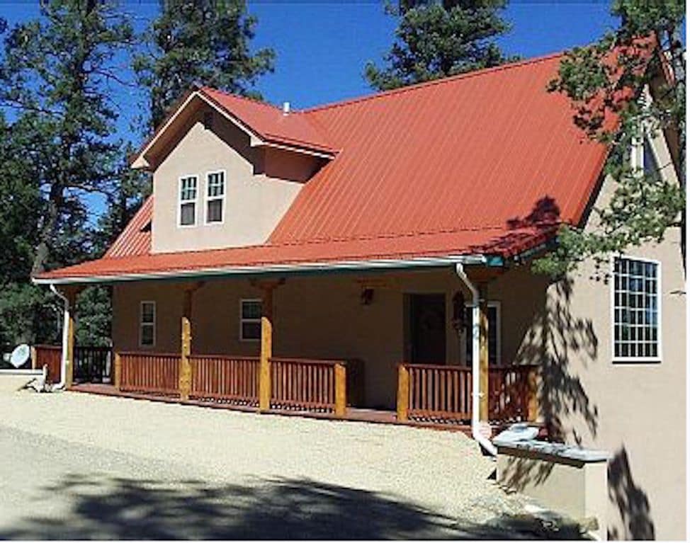 A cabin with a red metal roof is nestled among trees, featuring a covered porch with wooden railings. Large windows allow natural light, and a gravel pathway leads to the entrance. The surrounding area is dotted with greenery, enhancing its tranquil setting.