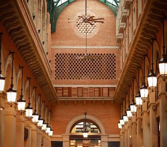 An indoor corridor is displayed, featuring an arched ceiling supported by colonnades. The warm tones of exposed brick contrast with the modern lighting fixtures. Rows of ornamental lanterns line the walkway, creating a welcoming ambiance while allowing natural light to filter through the upper glass structure.
