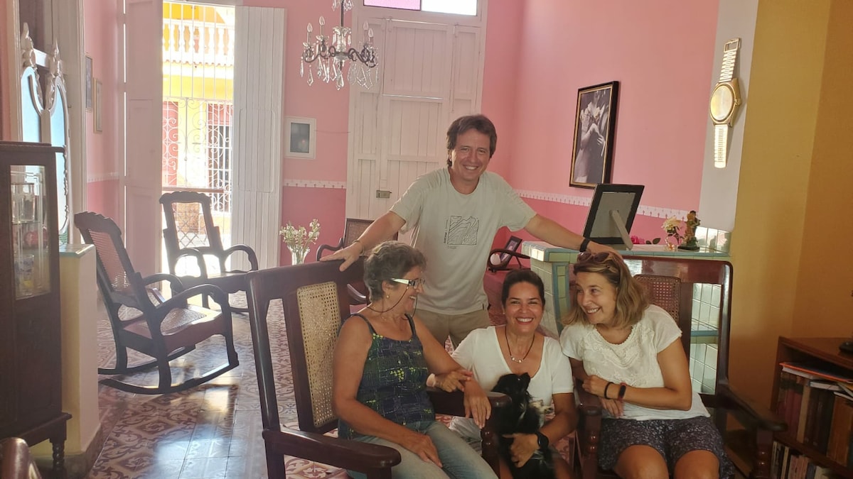 A welcoming living area features a blend of traditional and modern design. Comfortable rocking chairs are arranged around a central space. A vibrant pink wall enhances the ambiance, while a chandelier adds elegance. Natural light floods the area, creating a warm environment for gathering.