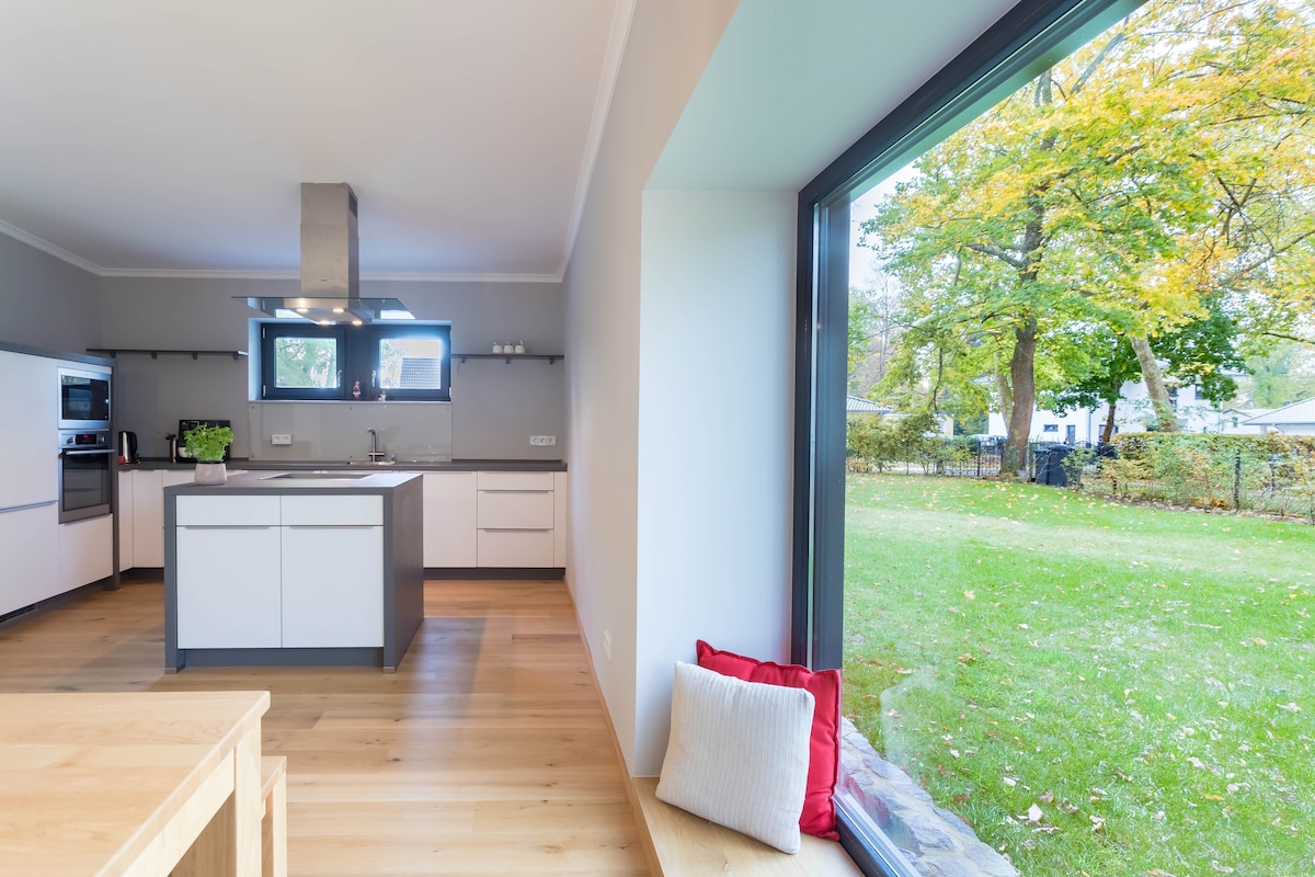 A modern kitchen is highlighted, featuring a central island and sleek cabinetry. Large windows provide a view of the well-maintained garden, allowing natural light to fill the space. An inviting nook with cushions is positioned by the window for relaxation.
