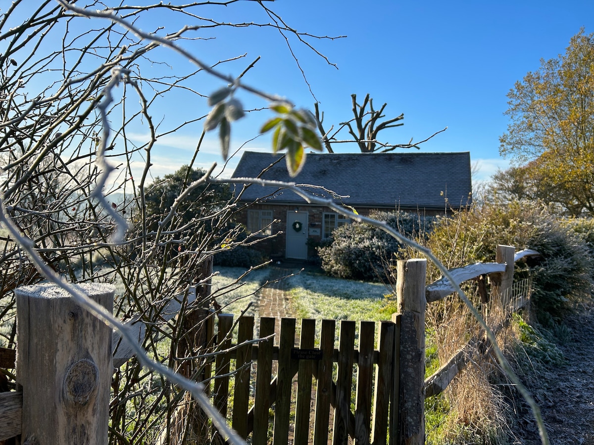 The Piggery is seen through a frosty wooden fence and branches, showcasing its rustic charm. Sunlight illuminates the exterior, highlighting the peaceful countryside setting. The inviting front door is framed by a wreath, with a backdrop of clear blue sky and a few trees in view.