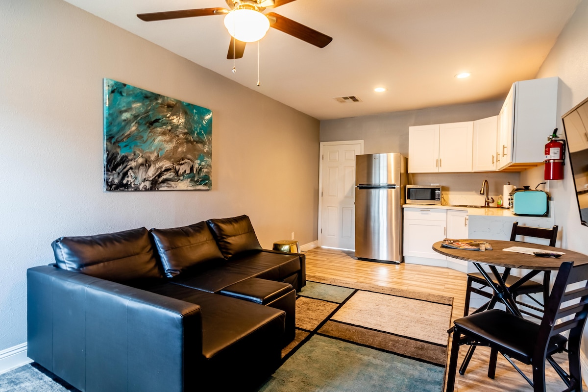 A modern living area features a sleek black couch positioned beside a small round dining table. The kitchenette is equipped with a stainless steel refrigerator and microwave. A colorful artwork adorns the wall, complemented by soft lighting from a ceiling fan.