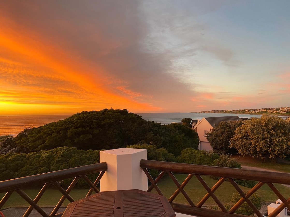 A captivating sunset paints the sky in vibrant oranges and yellows, as it sets over a lush landscape leading to the ocean. The view is framed by greenery, with the horizon showing gentle waves and distant coastal homes.
