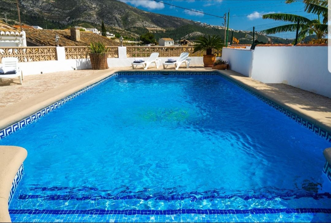 A private swimming pool is surrounded by a tiled terrace, featuring several sun loungers positioned for relaxation. Lush greenery is present nearby, with a scenic view of the mountains in the background and a clear blue sky overhead.