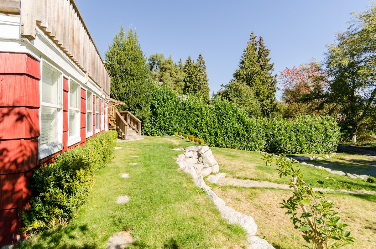 The exterior showcases a charming red house, surrounded by lush greenery. A stone pathway leads to a wooden staircase, inviting guests to explore. Vibrant grass contrasts with the carefully manicured hedges and trees, creating an inviting outdoor space.