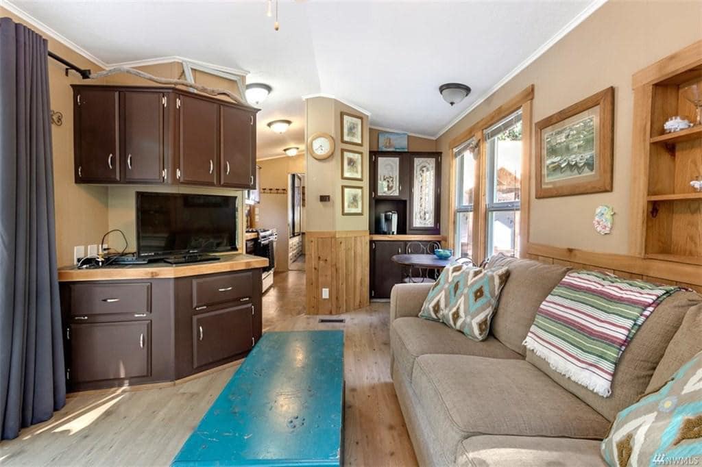 An open living area features a comfortable couch adorned with patterned pillows. A turquoise coffee table sits in front, with a wall of windows allowing natural light to illuminate the space. The kitchen is visible in the background, showcasing dark cabinetry and wall decor.