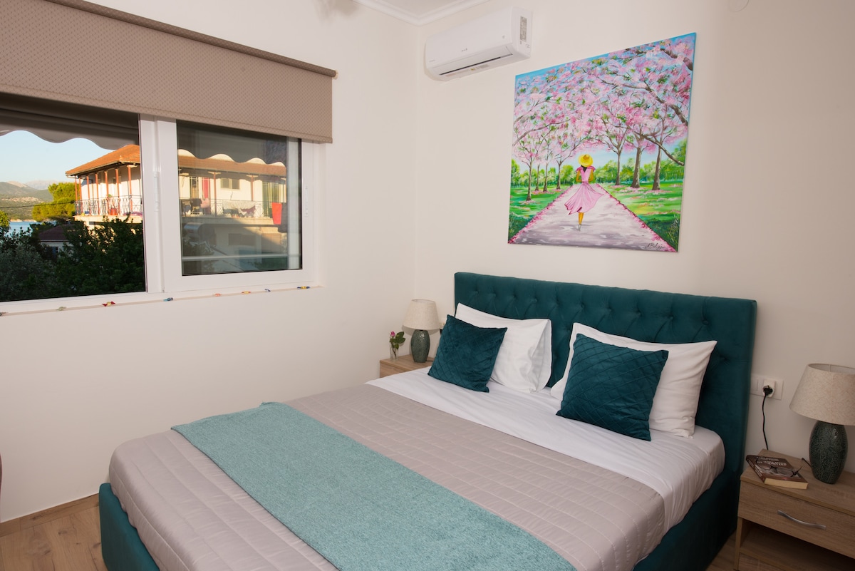 A spacious sleeping area features a teal upholstered bed with white and teal accent pillows. A light gray blanket is draped neatly over the bed. A floral artwork depicts a walking path under blooming trees, while natural light filters through a window adorned with shades.