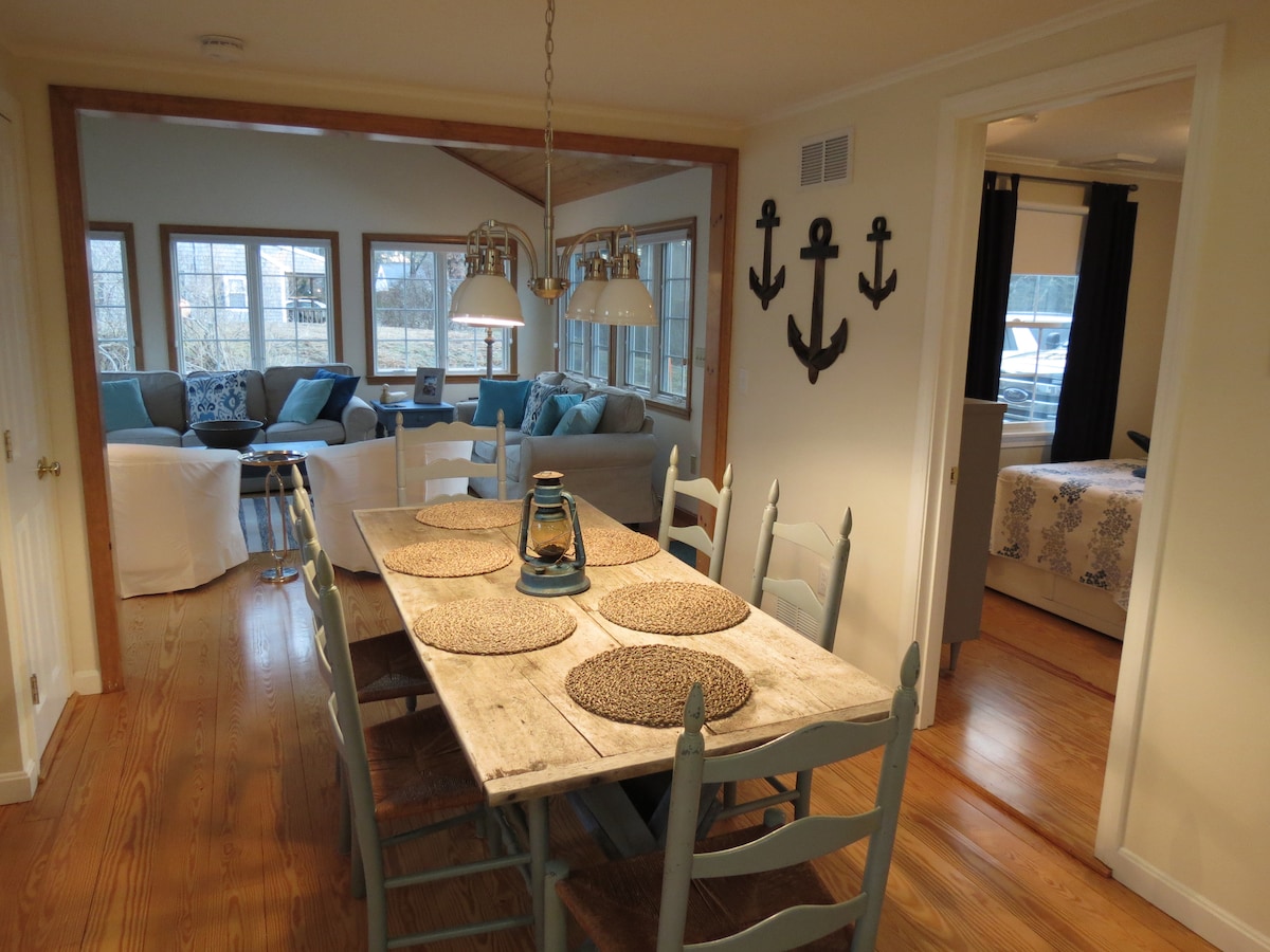 A rustic dining table is set with woven placemats, surrounded by light blue chairs. A lantern adds a warm touch. Beyond, a bright living area is visible, featuring a couch adorned with blue cushions, alongside large windows that allow ample natural light.