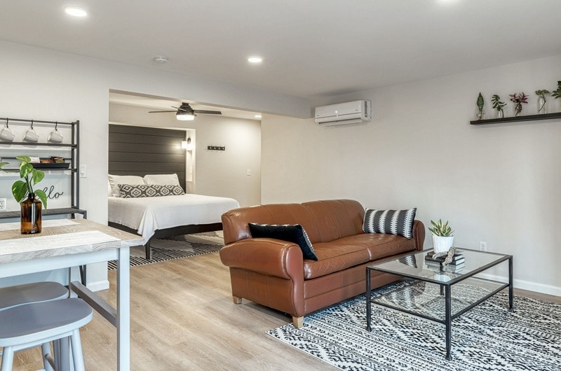 A modern studio space features a comfortable leather sofa positioned in front of a glass coffee table. A neutral color palette and light wood flooring are complemented by a decorative area rug. The sleeping area is visible, showcasing a spacious bed adorned with patterned bedding.