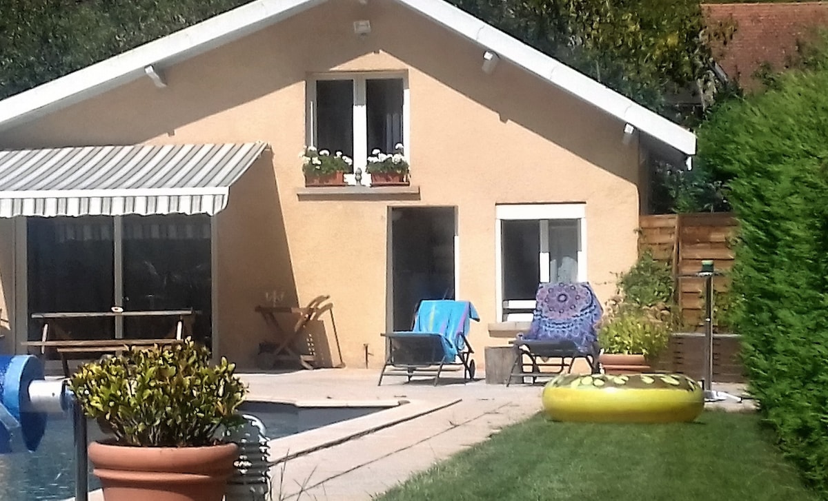 The exterior of a cozy house is visible, featuring a sunny patio with a striped awning. Two chairs are positioned nearby, alongside a round table. A yellow inflatable float and well-maintained greenery are seen by the pool, indicating a welcoming outdoor space.