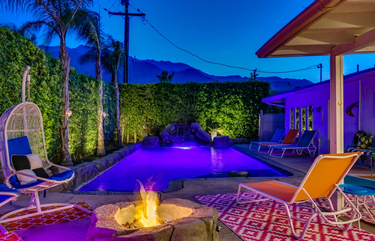 The outdoor area features a saltwater pool illuminated with soft purple lighting. Comfortable lounge chairs are arranged around the pool, and a fire pit sits on a patterned rug, providing a cozy gathering spot. Green hedges and palm trees enhance the secluded ambiance.