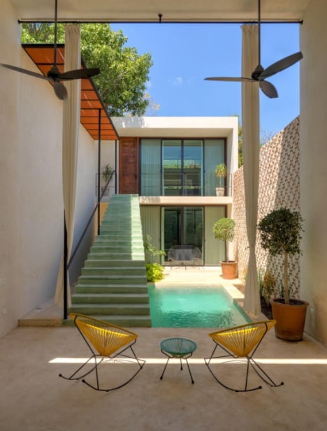 A serene private pool area is framed by modern architectural design, featuring a muted color palette. Two yellow lounge chairs are arranged around a small table, while lush greenery surrounds the pool, and a staircase leads to the upper level.