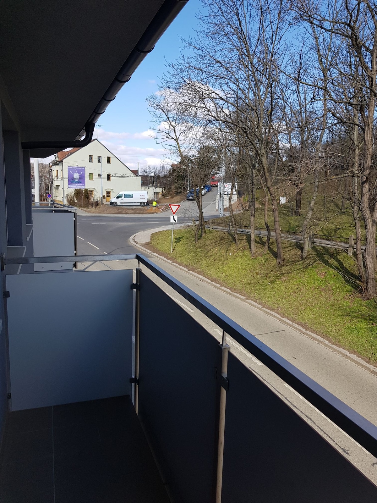 A balcony view captures a quiet street scene with a gentle curve. Surrounding greenery and a mix of residential buildings are visible. The sky is clear with a few clouds, enhancing the peaceful atmosphere of the location.