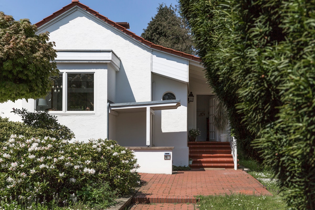 A charming exterior view of a white house, featuring a sloped red roof and a covered entryway. Lush greenery and flowering shrubs surround the entrance, which is approached by a brick pathway. A sunny blue sky provides a bright backdrop to this inviting façade.