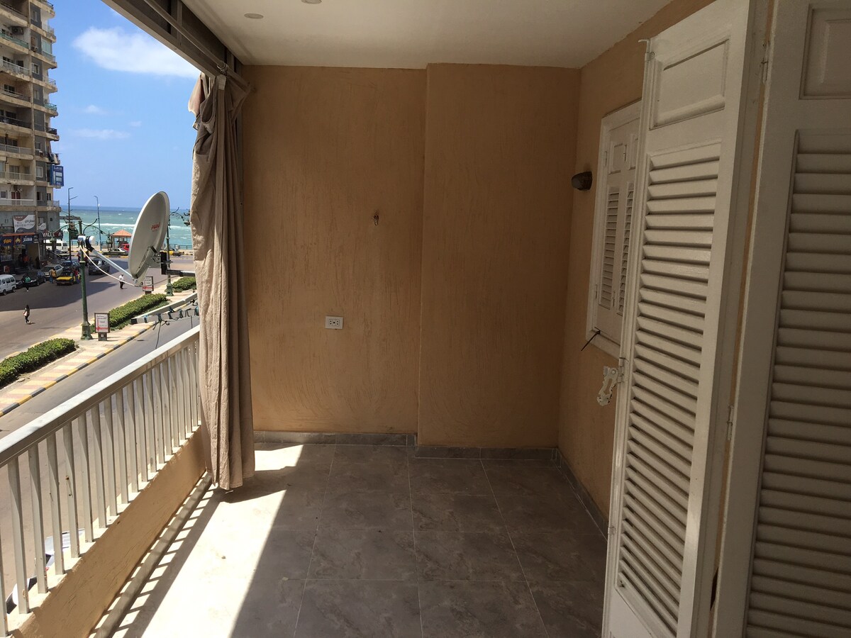 A spacious balcony features light-colored tiled flooring and beige walls, with partial views of the surrounding area. Light drapes frame the entrance, allowing natural light to fill the space. The scene outside includes a glimpse of the road and nearby buildings, inviting a refreshing sea breeze.