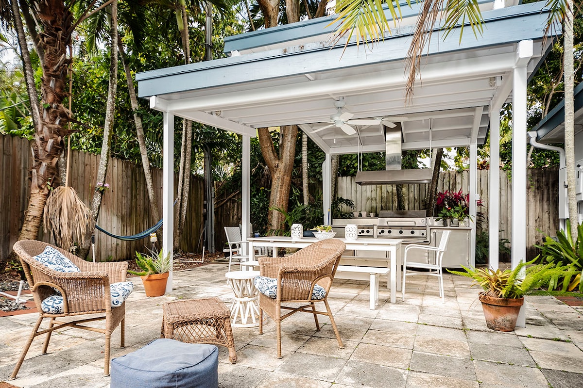 An outdoor kitchen area is featured under a spacious covered patio, equipped with an inviting grill. Comfortable seating arrangements include four chairs and a table, surrounded by lush greenery and tropical plants. A hammock is positioned nearby, enhancing the relaxed ambiance of the space.