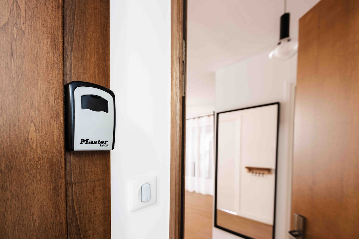 The entryway is equipped with a secure key box mounted on the wall, providing convenient access to the apartment. A light switch is visible, adding functionality. Directly across, a large mirror reflects the welcoming space, while a wooden door leads further inside.