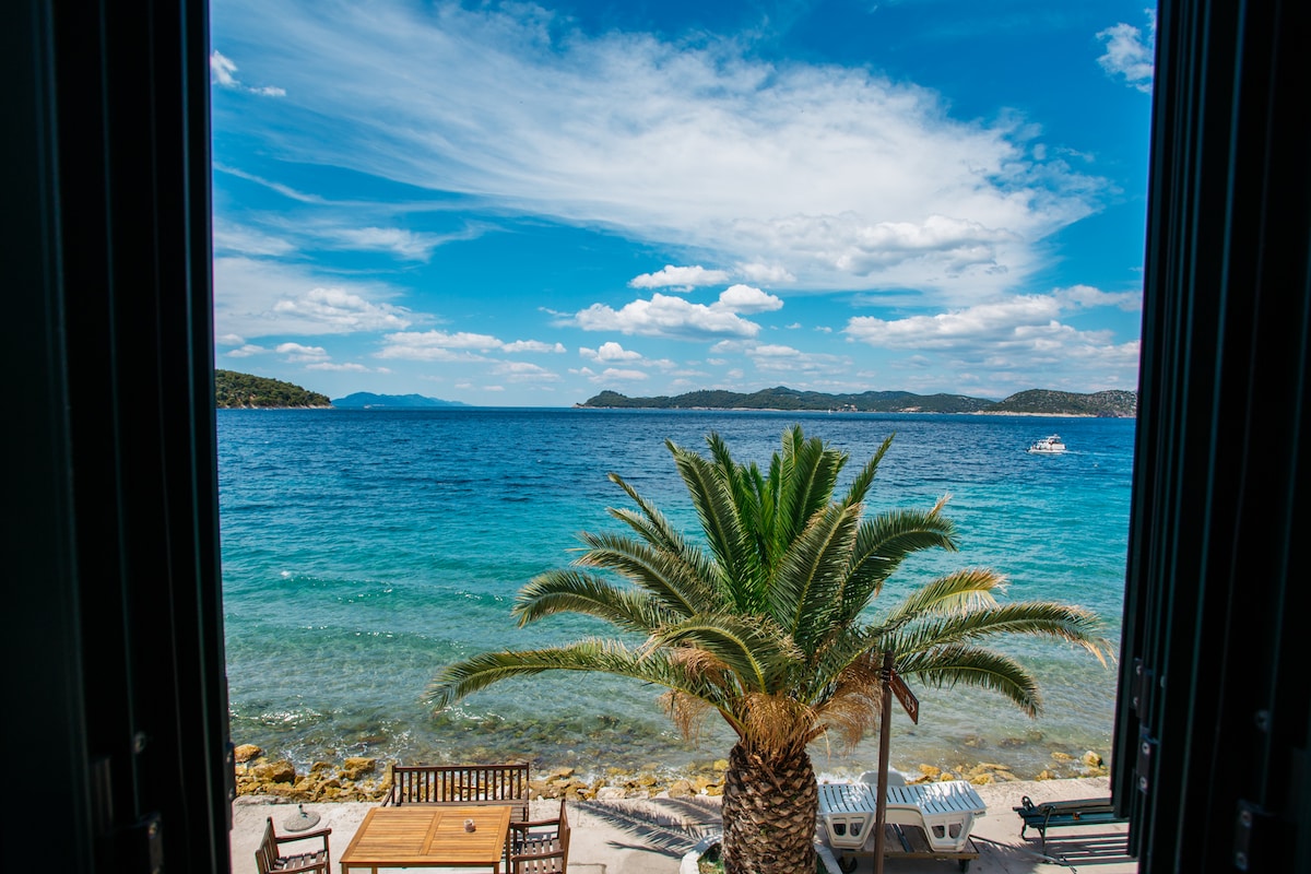 A view is captured from the apartment, showcasing the tranquil sea and distant islands under a blue sky. In the foreground, a palm tree stands next to wooden furniture arranged on the shore, inviting relaxation beside the water.