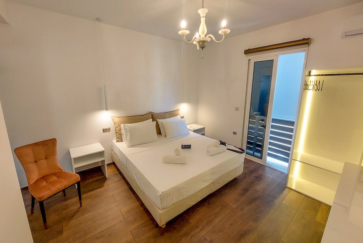 The open plan bedroom features a spacious double king size bed with light linens and two bedside tables. A chair is positioned in the corner, and sliding patio doors provide access to a small balcony, allowing natural light to fill the room.