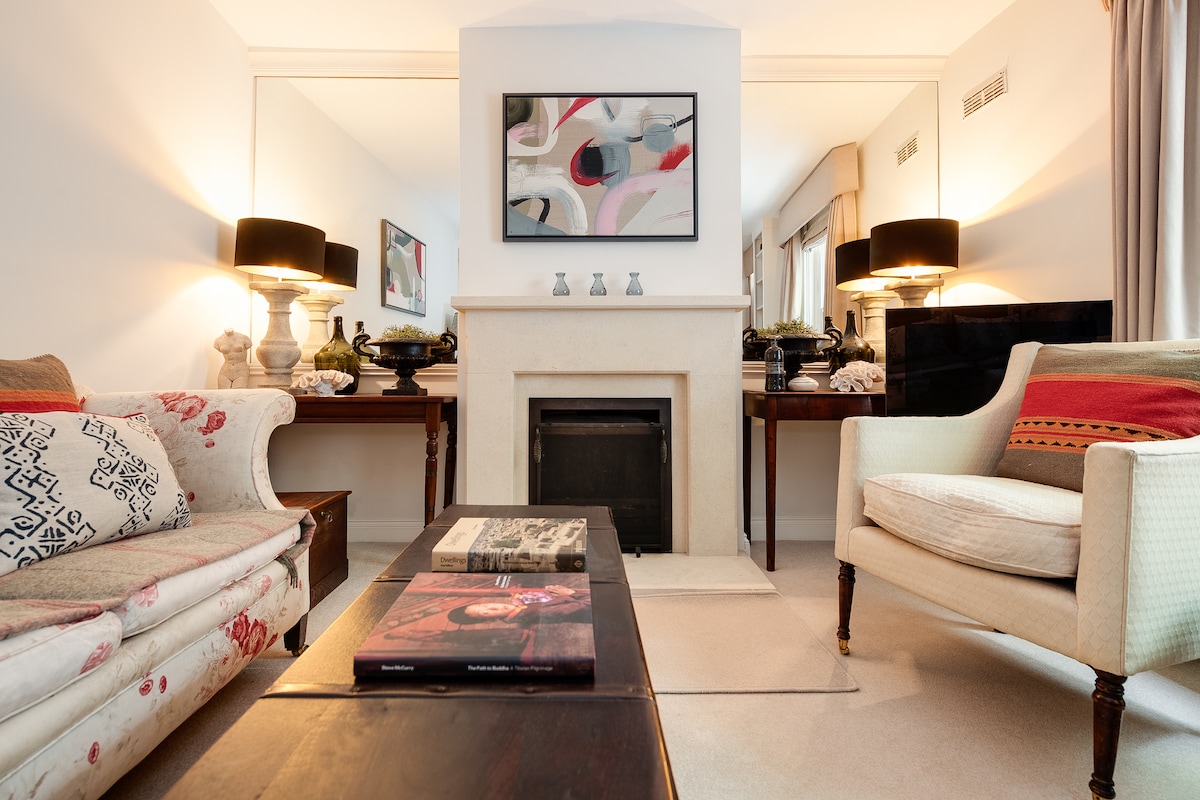 A lounge area reveals a fireplace framed by decorative mirrors. Sofas with patterned upholstery and solid armchairs create a cozy seating arrangement. A wooden coffee table holds stacked magazines, while lamps provide soft lighting. Artwork adorns the wall, adding color to the neutral palette.