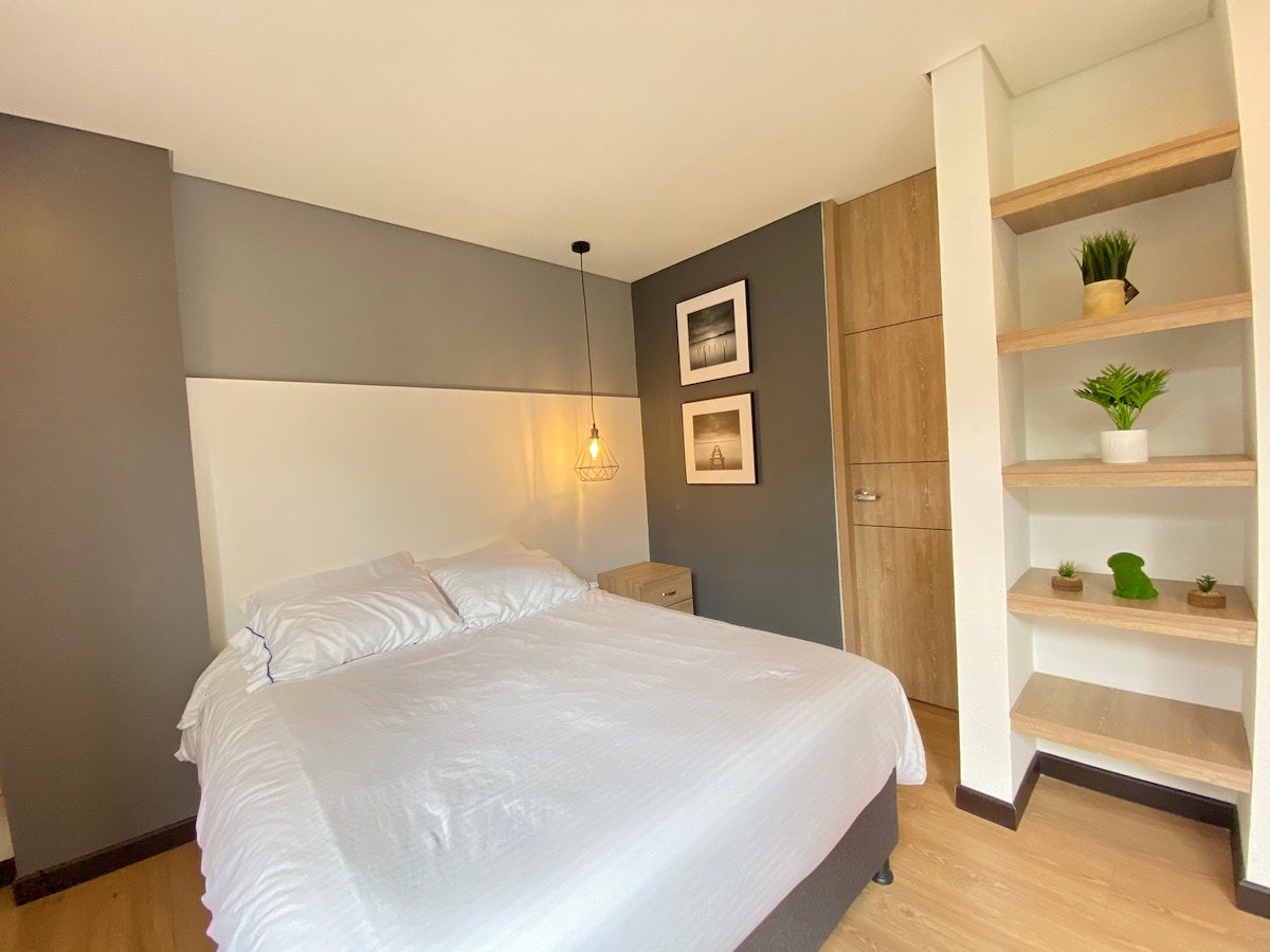 A cozy bedroom features a spacious bed with white linens and a headboard against a gray wall. A pendant light hangs above, while framed photographs are displayed on the wall. A nightstand and a built-in closet provide function, complemented by open shelving with plants.