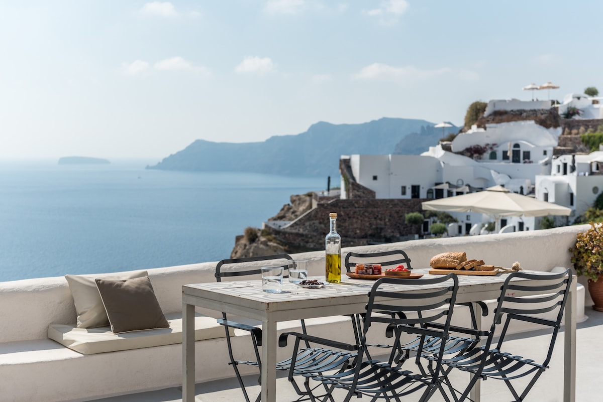 A dining table with six black metal chairs is positioned on a terrace overlooking the caldera. A bottle of olive oil, glasses, and freshly baked bread with pastries are arranged on the table. The vast blue sea and whitewashed buildings in the distance create a serene backdrop.