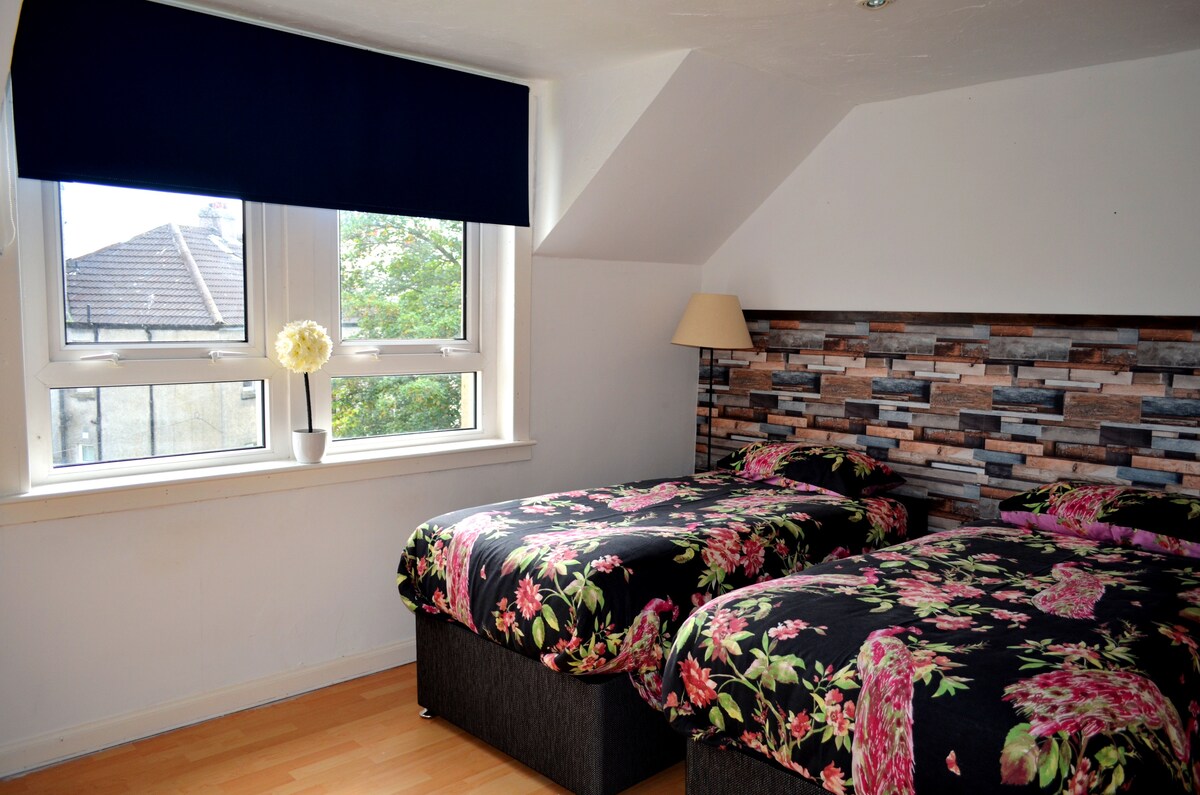 A twin bedroom features two beds with floral patterned bedding. A custom stone wall adds visual interest behind the beds. Natural light enters through two windows covered by a dark blind. A floor lamp provides additional lighting in the room.