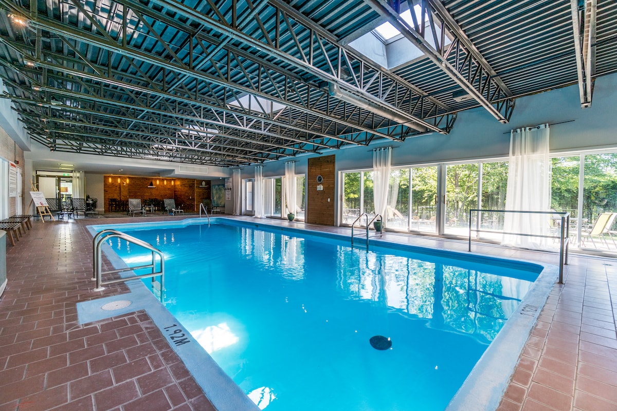 An indoor pool is situated under a high, textured ceiling with exposed beams. Sunlight filters through large windows, illuminating the clear blue water. Lounge chairs are arranged along the edges, and curtain panels allow for privacy while enhancing the bright ambiance.