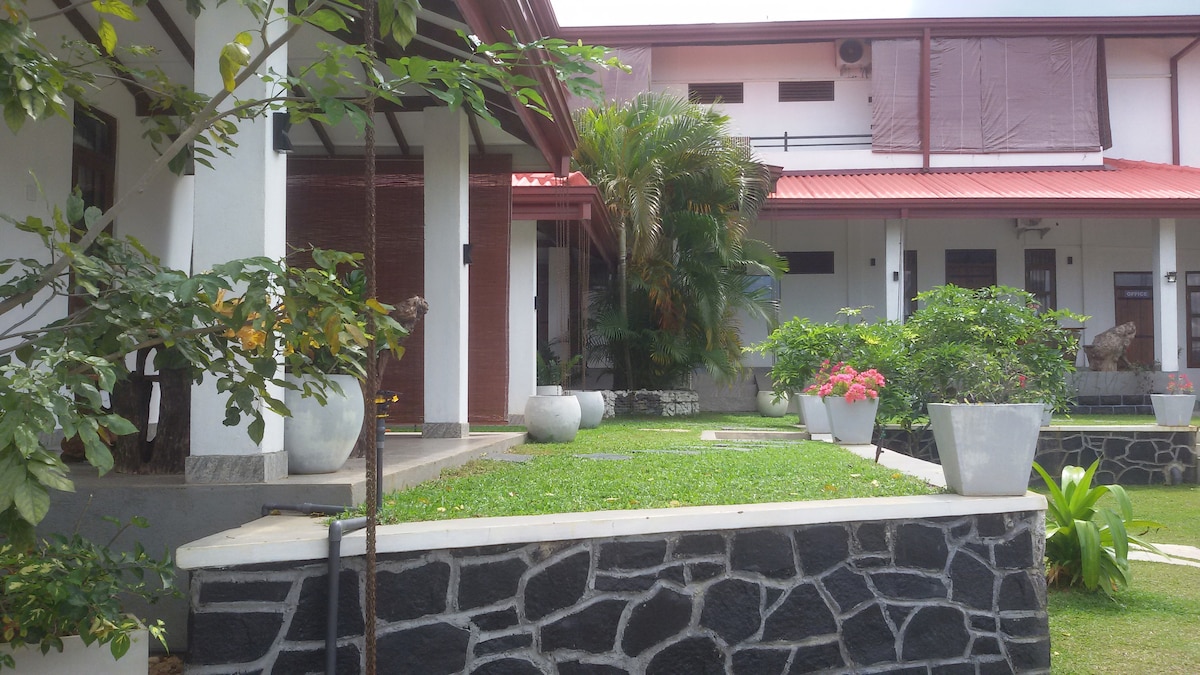 A serene garden area is depicted, featuring well-maintained green grass and potted plants. The building has a modern architectural style with a red roof and white walls. Pathways lead to various entrances, surrounded by lush foliage and decorative stones.
