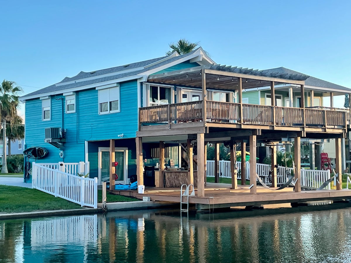 A vibrant blue waterfront house is displayed alongside a canal. The structure features multiple outdoor decks and a boat dock, providing easy water access. A spacious outdoor kitchen area is visible beneath the deck, enhancing the outdoor living experience.