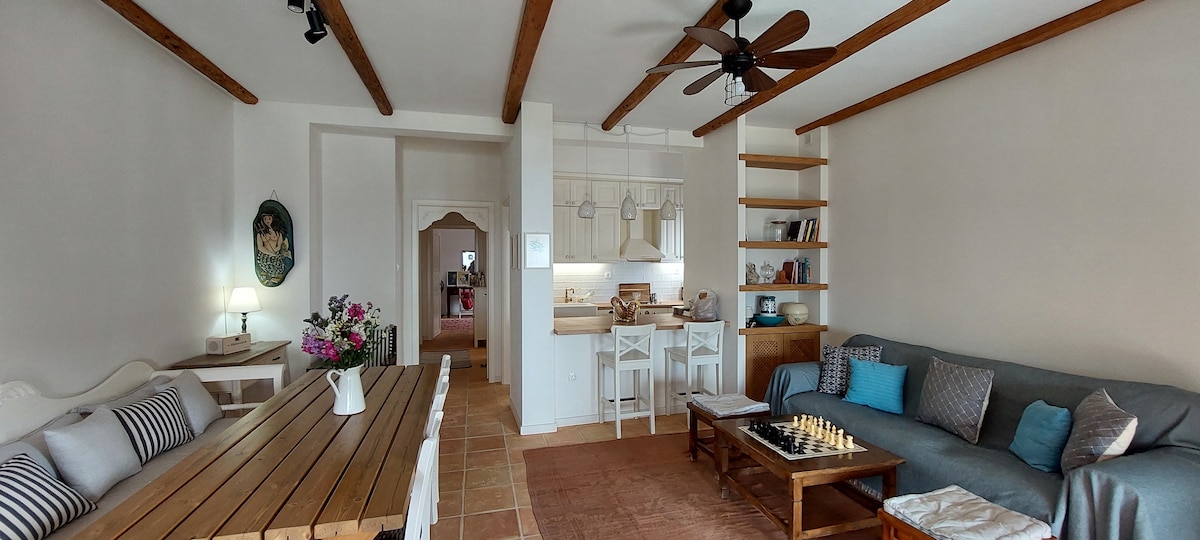 A spacious living area is highlighted by wooden beams overhead. A comfortable sofa faces a low coffee table with a chessboard arrangement. A dining table with rustic charm is positioned nearby, while an open kitchen area is visible in the background.