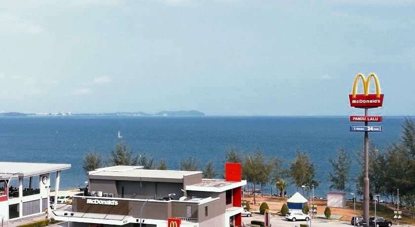 A coastal view showcases a clear blue ocean under a light sky. A McDonald's restaurant is visible in the foreground, with a prominent sign displaying the logo. Several trees line the shore, adding greenery to the scene.
