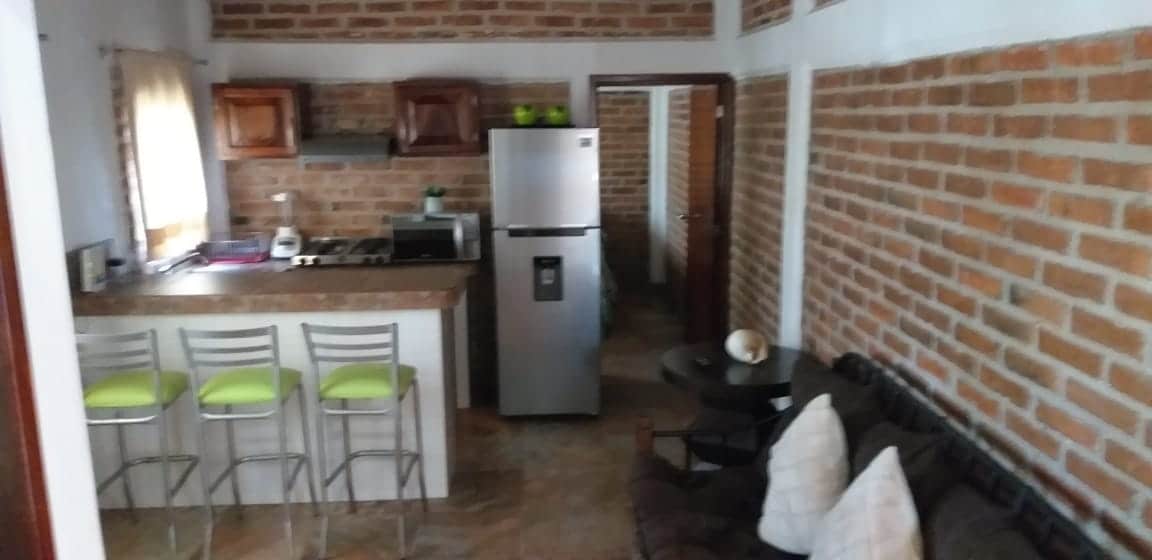 The open kitchen features exposed brick walls and a breakfast bar with four high stools. Stainless steel appliances are situated next to wooden cabinetry. A small seating area includes a dark sofa with decorative pillows, creating a cozy space for relaxation.